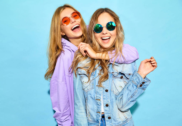 Two young beautiful blond smiling hipster girls in trendy summer clothes. Sexy carefree women posing near blue wall in sunglasses. Positive models going crazy - Foto, Imagem