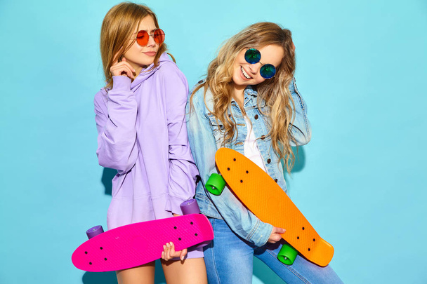 Two young stylish smiling blond women with penny skateboards. Girls in summer hipster sport clothes posing near blue wall in studio. Positive models - Fotografie, Obrázek
