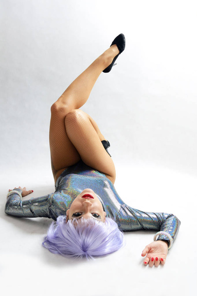 Jovencita, bailarina disfrazada. Modelo femenino con traje metálico especial y peluca púrpura clara. Chica tendida en el suelo con las piernas arriba
. - Foto, imagen