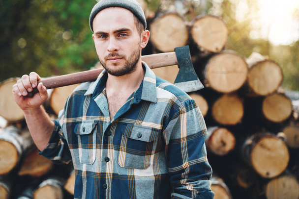 Strong bearded lumberman holds ax on his shoulder on warehouse of logs. Blurred background - Valokuva, kuva