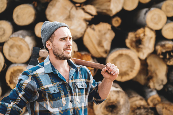 Strong bearded lumberjack wearing plaid shirt hold ax in hand on background of sawmill and warehouse of trees - Valokuva, kuva