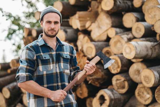Bearded lumberjack wearing plaid shirt hold ax in hand on background of sawmill - Photo, image