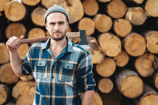 Strong bearded lumberman wearing plaid shirt holds axe on his shoulder on background of sawmill - Foto, imagen