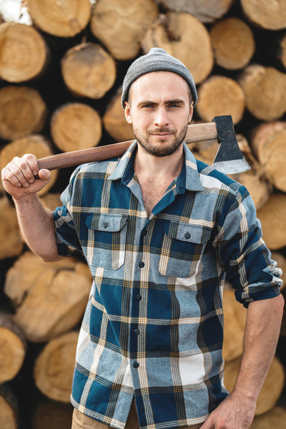 Strong bearded woodcutter holds axe on his shoulder on warehouse of logs. Blurred background - Zdjęcie, obraz