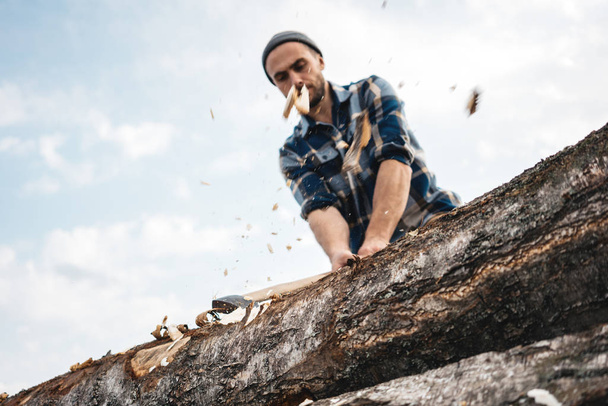 Strong and brutal lumberjack with ax in his hands chops tree in forest, wood chips fly apart. Blurred background - Photo, image