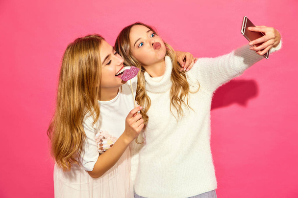 Portrait of two young stylish smiling blond women. Girls dressed in summer hipster clothes. Positive models making selfie on smartphone on pink background - Fotó, kép