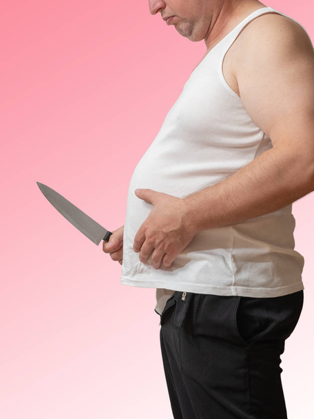 Een man met één hand houdt dat de vet Vouw op zijn grote buik en in de andere hand houdt een mes, alsof willen verwijderen. - Foto, afbeelding