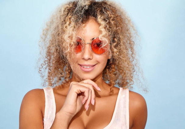 Portrait of blissful lovable woman with blond african hairstyle. Smiling model girl in trendy summer clothes posing on blue background in sunglasses - Φωτογραφία, εικόνα