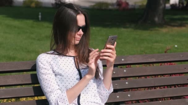 Young woman brunette types a message on mobile phone sitting on the bench in park. - Séquence, vidéo