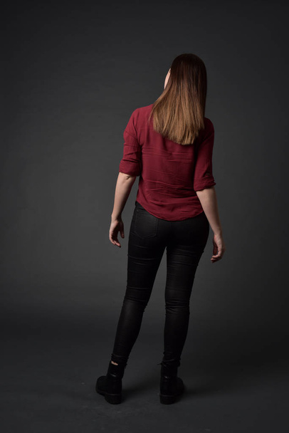 retrato de longitud completa de chica morena con camisa roja y pantalones de cuero. pose de pie, sobre fondo gris estudio
. - Foto, Imagen