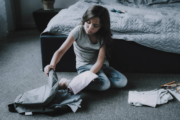 Cute Girl Packing Bag Sits on Floor in Bedroom. Serious Black-Haired Caucasian Child Wearing Blue Jeans and Gray T-Shirt Sitting on Gray Carpet Near Large Bed. Kid Picking School Supplies - Foto, Imagem