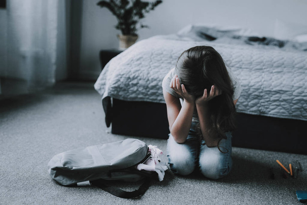 Girl Sitting on Floor in Bedroom and Crying. Portrait of Unhappy Black-Haired Child with Closed Eyes Covers Face with Hands Wearing Gray T-Shirt Sits Near Backpack next to Bed in Modern Apartment - Фото, изображение