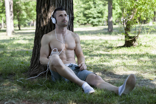 Hezký a atraktivní svalnatý mladý muž, shirtless v parku a poslechu hudby. Ruka držící mobilní telefon. - Fotografie, Obrázek