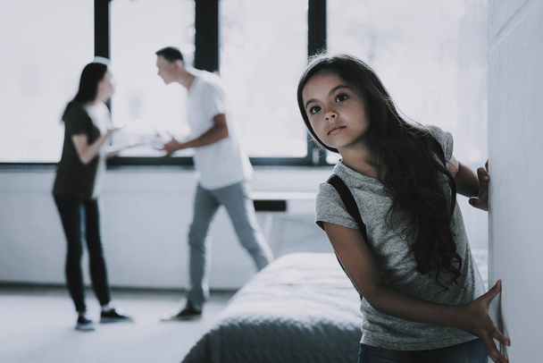 Dark Haired Girl Listens Parents Arguing at Home. Portrait of Child Looks Gloomy Leaning Against Wall While Mother and Father Scolding in Living Room. Family Relationship Problems Concept - Foto, Imagen