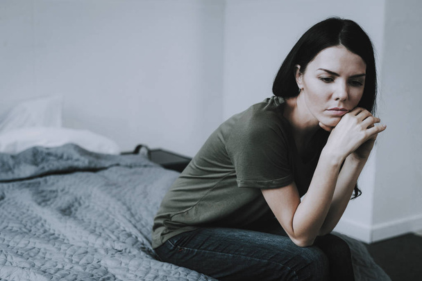 Portrait of Sad Dark-Haired Woman Sits on Bed. Gloomy Caucasian Frowning Person Wearing T-Shirt and Jeans Keeps Head on Hands Sits on Plaid in Bedroom with Light Gray Backround - Foto, imagen