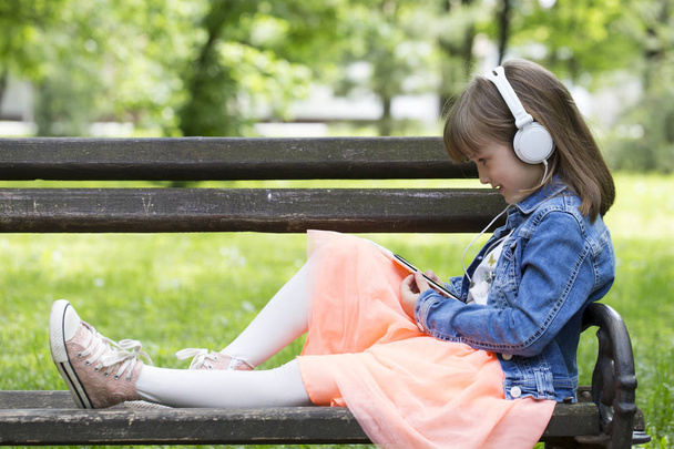 Klein meisje in een rok die zittend op een bankje, luisteren naar muziek en kijken naar mobiele telefoon - Foto, afbeelding
