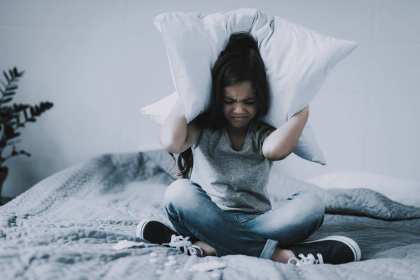 Girl Sitting Cross-Legged Covers Head with Pillow. Closeup Portrait of Frowning Black-Haired Caucasian Little Child Wearing T-Shirt and Jeans Sits on Gray Bed Closes Eyes and Ears. - Фото, зображення