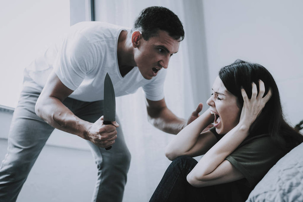 Angry Man Holds Knife and Screams at Wife at Home. Woman Sitting on Floor Near Gray Bed Clasps Head Cries in Fear. Married Couple Argues in Bright Bedroom. Quarrel and Family Violence Concept - 写真・画像