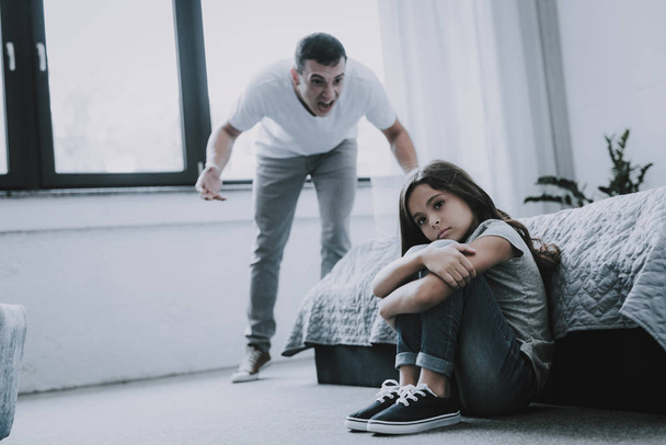 Man Screams at Sad Girl who Sitting on Floor. Indifferent Child Wearing Gray T-Shirt Turns Away Clasps Around Legs Looks Off to Side While Father Yelling. Child Offense Concept - Fotografie, Obrázek