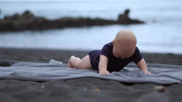 El bebé yace boca abajo sobre la arena negra cerca del océano y se ríe mirando a la cámara
 - Imágenes, Vídeo
