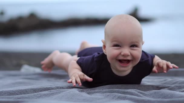 Dítě leží na břiše na černém písku nedaleko oceánu a směje se, při pohledu na fotoaparát - Záběry, video