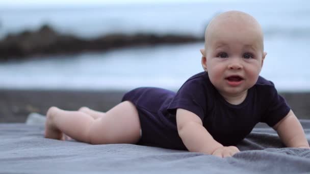 The baby lies on his stomach on the black sand near the ocean and laughs looking at the camera - Footage, Video