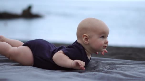 Дитина лежить на животі на чорному піску біля океану і сміється дивитися на камеру
 - Кадри, відео