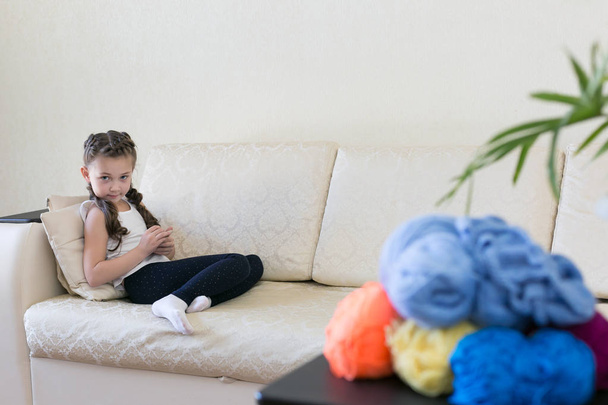 On the table there are threads for knitting in different colors. A girl with a mobile phone on the couch. - Φωτογραφία, εικόνα