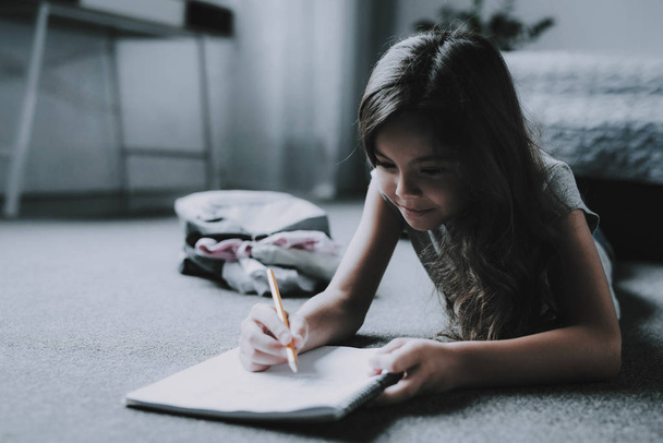 Cute Little Girl Draws in Notepad Lying on Floor. Portrait of Black-Haired Child Wearing Casual Clothes Lying on Gray Carpet and Writes with Pencil in White Notebook. Leisure Time Concept - Фото, изображение