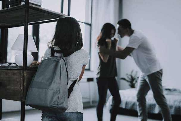 Sad Girl Watches How Parents Fighting at Home. Portrait of Child Wears Gray T-Shirt Stands Near Rack While Mother and Father Scolding in Living Room on Backround. Family Conflict Concept - Foto, Imagen