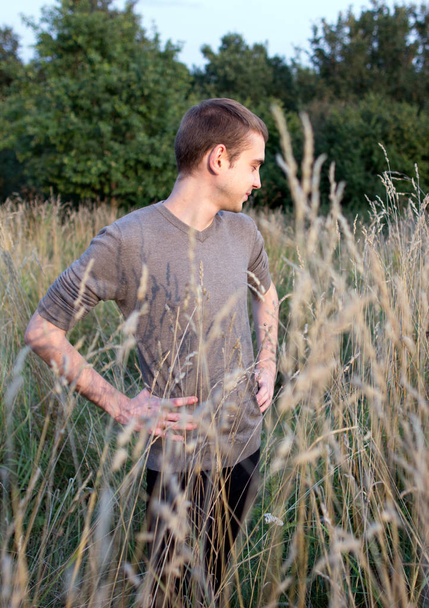 A young attractive thin guy standing in the tall grass on the forest background. A young student side view portrait in the park. - Fotoğraf, Görsel