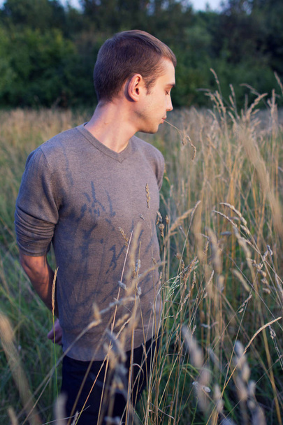 A young attractive thin guy standing in the tall grass on the forest background. A young student side view portrait in the park. - Fotoğraf, Görsel