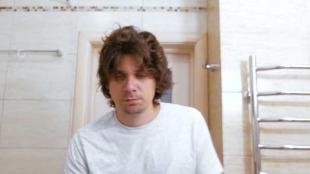 Sleepy shaggy young man looks at the mirror in bathroom in the morning and combs long hair with hands. - Filmagem, Vídeo