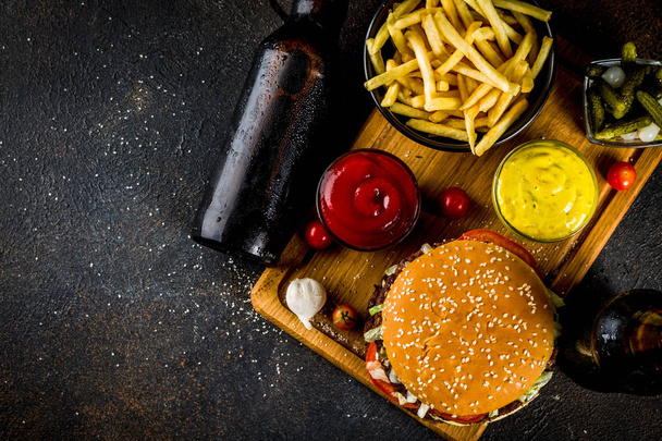 Various party food, Hamburgers, French fries, potato chips, pickled cucumbers, onions, tomatoes and cold beer bottles, rusty black concrete background copy space - Foto, Imagen