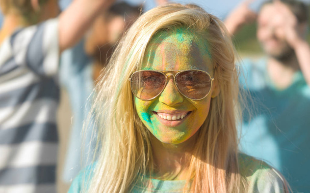 Portrét krásné blond Girl slaví Festival Holi se svými přáteli. Její tvář a oblečení jsou pokryty barevný prášek. Ona se dívá do kamery. - Fotografie, Obrázek