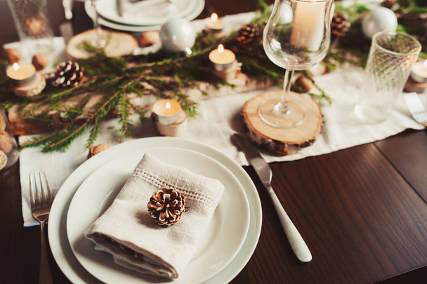 Feestelijke kerst- en nieuwjaarstafel in Scandinavische stijl met rustieke handgemaakte details in natuurlijke en witte tinten. Eetplaats versierd met dennenappels, takken en kaarsen - Foto, afbeelding