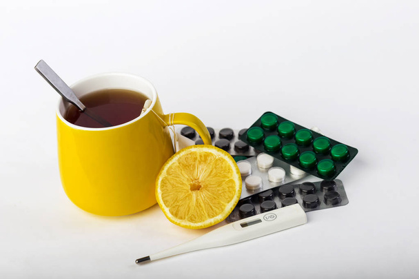Žlutý hrnek čaje s citronem, teploměrem a prášky na léčbu studenou a chřipku. Prevence nachlazení a chřipky na šedém pozadí - Fotografie, Obrázek