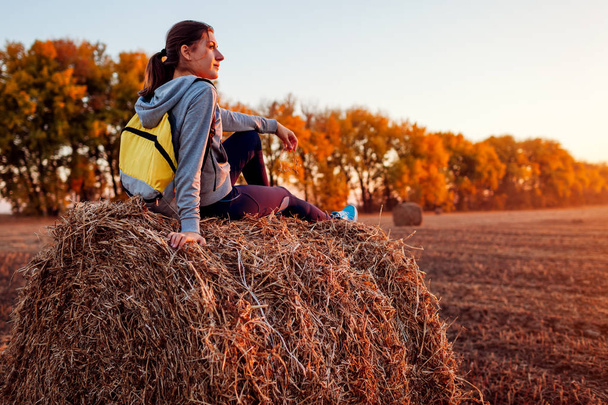 Young traveler having rest in autumn field at sunset. Woman admiring view sitting on haystack. Sport recreation concept - Foto, Imagem