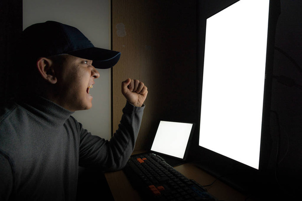 Vista lateral del hombre Hacker sentarse en el monitor de la computadora, tableta de pantalla blanca y enojado y molesto sentimiento en el cuarto oscuro
. - Foto, Imagen