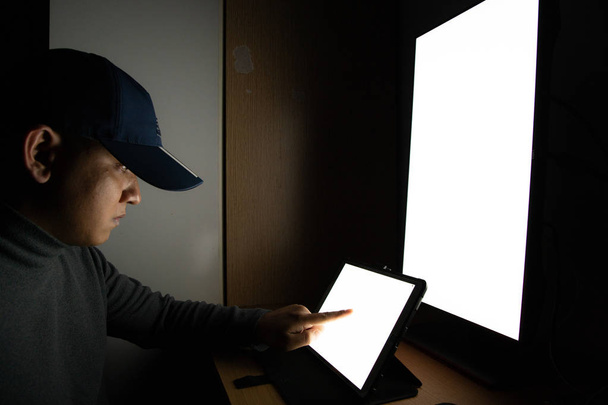 Vista lateral del hombre Hacker sentarse en el monitor de la computadora, tableta de pantalla blanca señalando el monitor de los dedos en la habitación oscura
. - Foto, Imagen