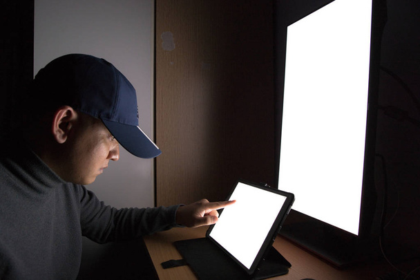Vista lateral del hombre Hacker sentarse en el monitor de la computadora, tableta de pantalla blanca señalando el monitor de los dedos en la habitación oscura
. - Foto, Imagen
