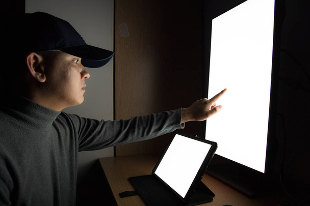 Boční pohled na člověka hacker sedí u monitoru počítače, bílá obrazovka tablet ukazující na prsty v tmavé místnosti. - Fotografie, Obrázek