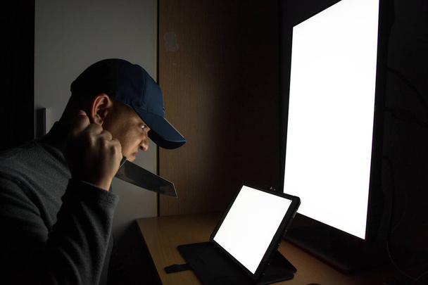 Boční pohled na člověka hacker sedí u počítačového monitoru, bílá obrazovka a drží nůž. v temné místnosti. - Fotografie, Obrázek
