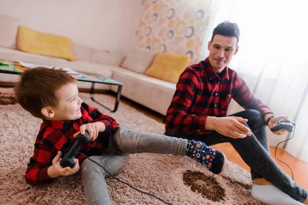веселый взволнованный отец и маленький сын в красных рубашках играют в консольную игру с геймпадами
 - Фото, изображение