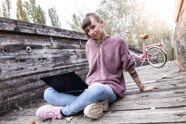 молода жінка навчається з комп'ютером, сидячи на дерев'яному мосту в парку серед осіннього листя
 - Фото, зображення