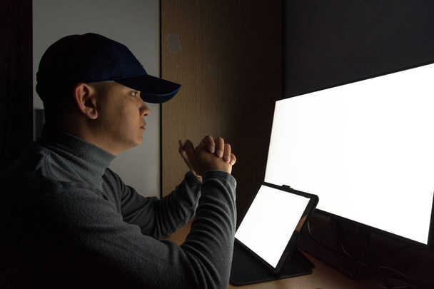 Vista lateral do homem Hacker sentar-se no monitor do computador, tablet tela branca no quarto escuro
. - Foto, Imagem