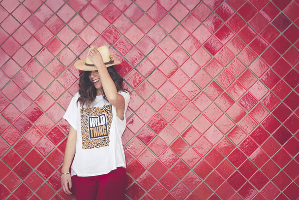 Chica posando con un sombrero delante de una pared roja. Ella tiene el sombrero en la mano. Lleva pantalones rojos y una camiseta blanca. Foto horizontal con espacio a la derecha
 - Foto, imagen