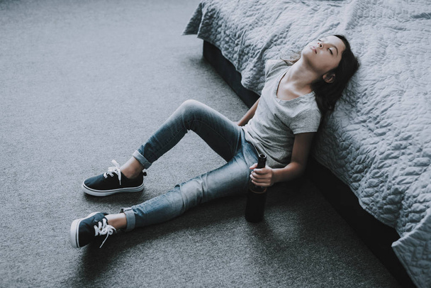 Drunk Sleeping Little Girl Sits on Floor near Bed. Frowning Unhappy Black-Haired Caucasian Child Wearing Gray T-Shirt Holds Alcohol Drink in Hand Leans on Bed Edge. Children Alcoholism Concept - 写真・画像