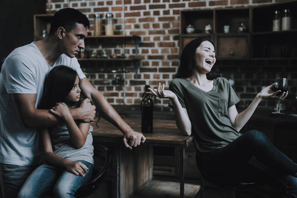 Father and Child Look at Drunk Mother at Kitchen. Woman Sits on Chair in Modern Room in Wooden Style Hold Glass of Wine in Hand and Laughs Loudly. Husband Hugs and Protects Kid. Alcoholism Concept - Foto, Imagem