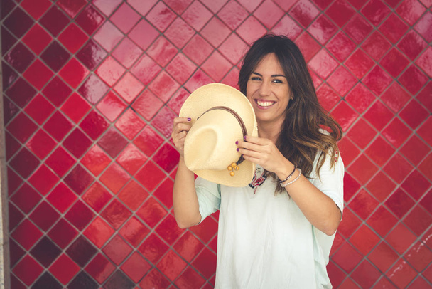 Девушка позирует в шляпе перед красной стеной. У неё в руке шляпа. Она носит красные брюки и белую футболку. Горизонтальное фото с пространством справа
 - Фото, изображение